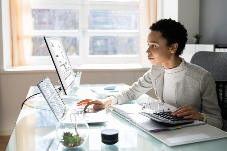 A person sits at a desk, working on a laptop and desktop computer, providing IT support. They use a calculator with papers nearby. The office is bright, with large windows allowing natural light to stream in. A small plant adds a touch of greenery amidst the office items.