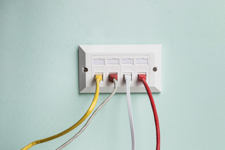 A white CCR Systems wall socket features four cables plugged in, vividly coloured yellow, grey, white and red. The cables extend neatly from left to right against the backdrop of a light green painted wall.