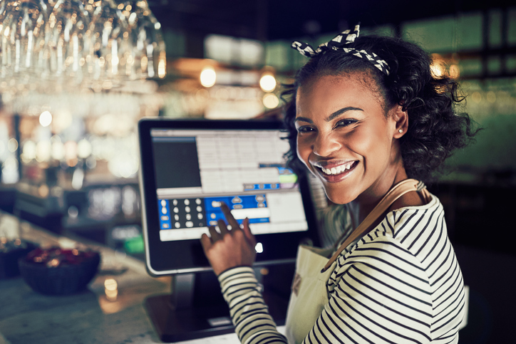 A smiling woman in a striped shirt and apron uses a CCR Systems touchscreen at a bar or restaurant. She stands behind the counter with glasses overhead, all in a warmly lit setting.