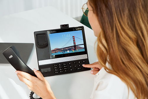 A person with long hair sits at a desk, using a CCR Systems touchscreen telephone displaying the Golden Gate Bridge. They hold the handset and press a button while a laptop is partially visible on the desk.
