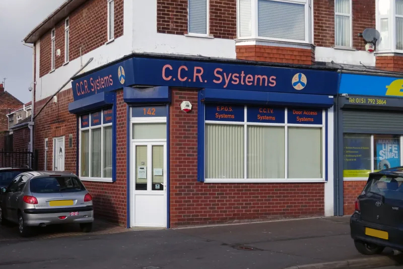 On a street corner, a brick building features blue signage for C.C.R. Systems, recently acquired by Excel Connect Solutions. The shopfront exhibits services such as EPOS Systems, CCTV Systems, and Door Access Systems. Two parked cars are visible on the road in front.