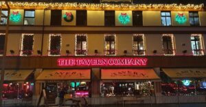 A street view of The Tavern Company at night, adorned with Christmas lights and wreaths. The neon sign glows pink above the entrance. Windows featuring holiday decor are visible, highlighting a successful festive atmosphere both inside and outside.