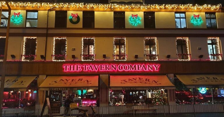A street view of The Tavern Company at night, adorned with Christmas lights and wreaths. The neon sign glows pink above the entrance. Windows featuring holiday decor are visible, highlighting a successful festive atmosphere both inside and outside.