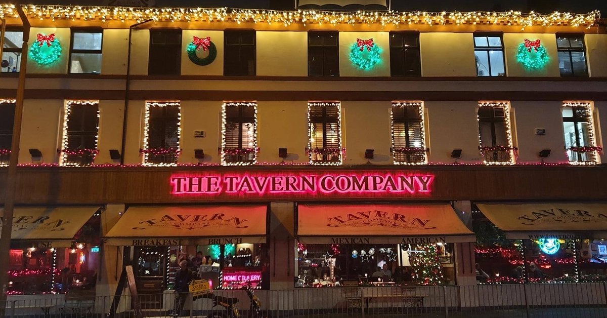 A street view of The Tavern Company at night, adorned with Christmas lights and wreaths. The neon sign glows pink above the entrance. Windows featuring holiday decor are visible, highlighting a successful festive atmosphere both inside and outside.