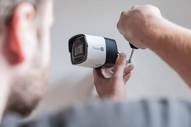 A person is installing a Panoramic QD security camera, part of the CCR Systems line, on a wall using a screwdriver. The sleek black-and-white camera complements the scene as the person’s hands expertly work, with part of their head visible in the foreground.