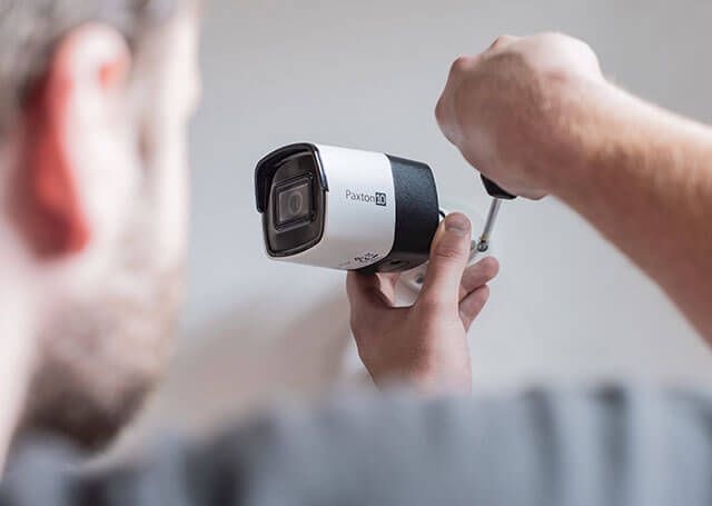 A person is fitting a security camera from CCR Systems onto a wall. The camera, mainly white and black, gleams under the light as the person skilfully uses a screwdriver to fix it in place. The emphasis is on the precision of the installation and the quality of CCR Systems design.