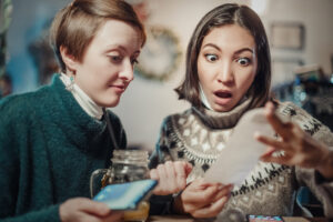 Two women sit together at a restaurant; one holds a smartphone and a Kilner jar, while the other looks surprised at a long receipt. Cosy jumpers drape them as they choose from an EPOS system, seated at a table with a beautifully blurred background.