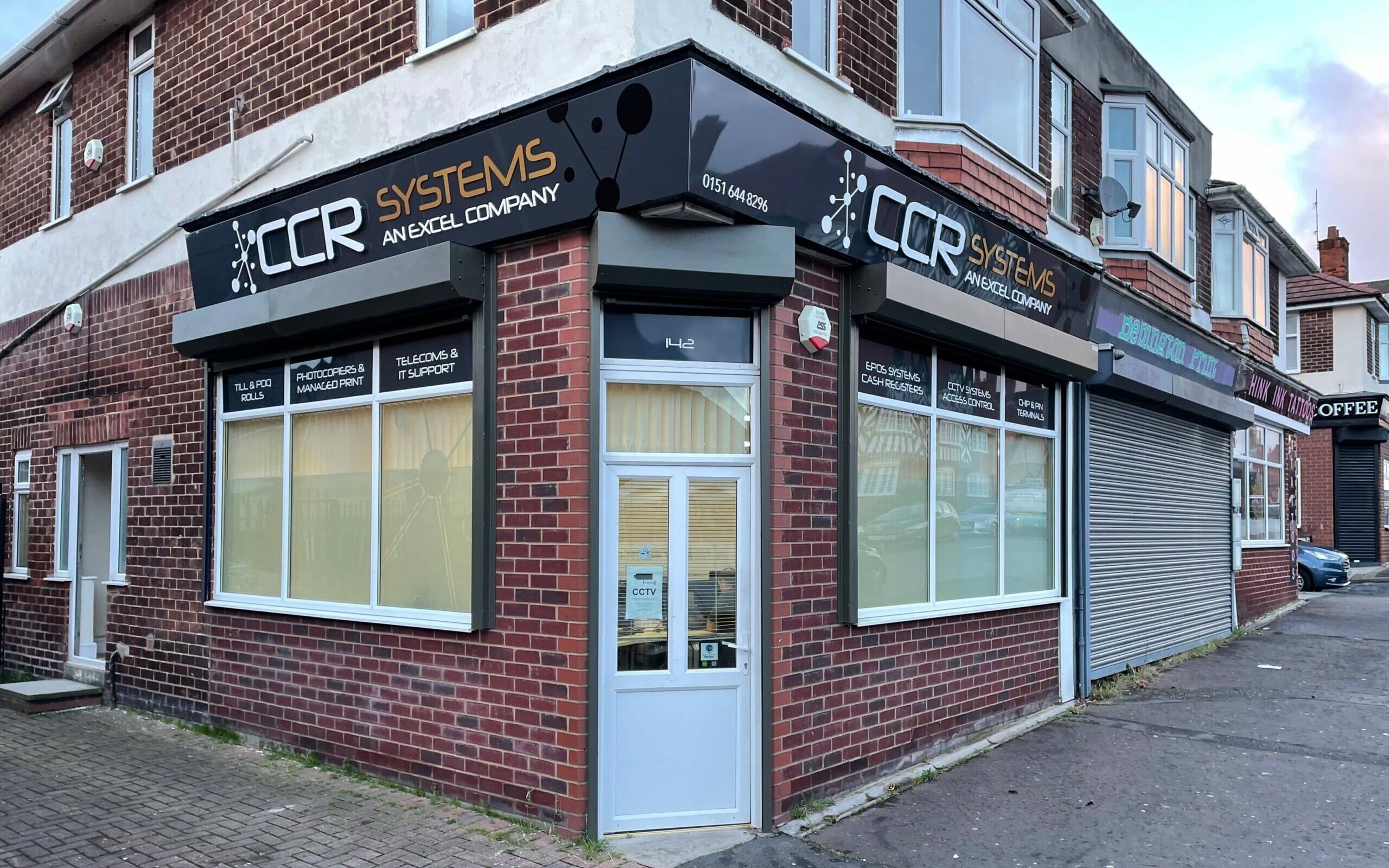 A picture of CCR Systems retail office in New Ferry, Wirral. The signage has a black background with white text and reads CCR Systems an Excel Company. The roller shutters have been painted black to match the signage. Above each of the windows, a different product is listed, ranging from cash registers to CCTV systems.