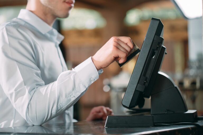 A person in a white dress shirt operates a touchscreen at an EPOS terminal on the counter, streamlining restaurant operations. The background is softly blurred, drawing attention to the activity at the device.