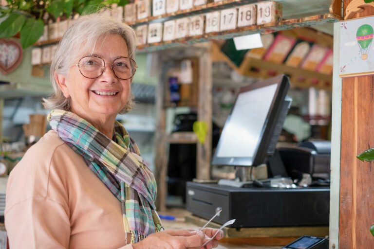An older woman with glasses and a scarf smiles warmly by the till in a cosy shop. She holds a receipt, demonstrating seamless payments through an EPOS system. In the background, a calendar and colourful decorations add to the welcoming atmosphere.