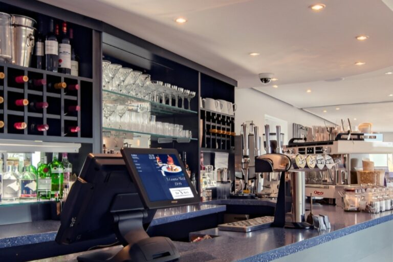 A modern bar interior featuring a counter with a sleek ICRTouch EPOS system, an assortment of glasses, bottles, and taps for serving drinks. Shelving holds various spirits bottles and wine glasses, with a stylish ceiling and recessed lighting above—a testament to innovative partnerships in design.