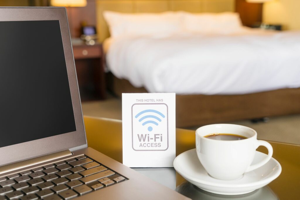 A sleek silver laptop sits on a glass table in a stylish hotel room. Next to it, a cup of coffee rests on a saucer, and beside it, a small sign highlights the available Wi-Fi access points. A neatly made bed with fluffy pillows is blurred in the background.
