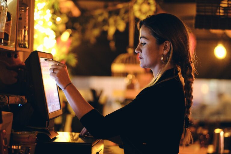 A woman with a plaited hairstyle is using a touchscreen in a dimly lit setting, possibly equipped with England EPOS Systems. She is focused on the screen, with warm lighting and blurred greenery in the background, suggesting she might be managing orders in a cosy café or bar.