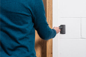 A person in a blue jumper uses a handheld device near a wooden door, employing an access control system. The device is held against a white brick wall, possibly as a security measure to safeguard assets and data.
