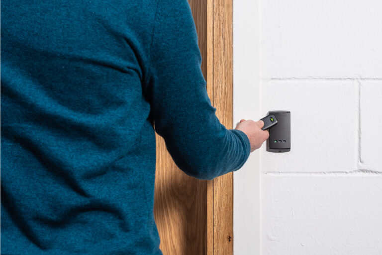 A person in a blue jumper uses a handheld device near a wooden door, employing an access control system. The device is held against a white brick wall, possibly as a security measure to safeguard assets and data.