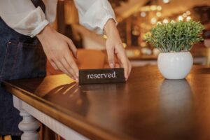 A persons hands place a reserved sign on a wooden table in a warmly lit restaurant. Thanks to the efficiency of online booking systems, even SMEs can manage reservations effortlessly. A small potted plant sits on the table, adding charm to the setting.