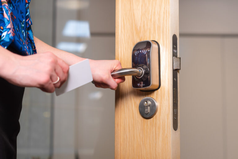 A person holds a keycard and uses it to unlock a door equipped with an access control system. The door is partially open, revealing a modern handle and keyhole. The person wears a blue floral shirt.