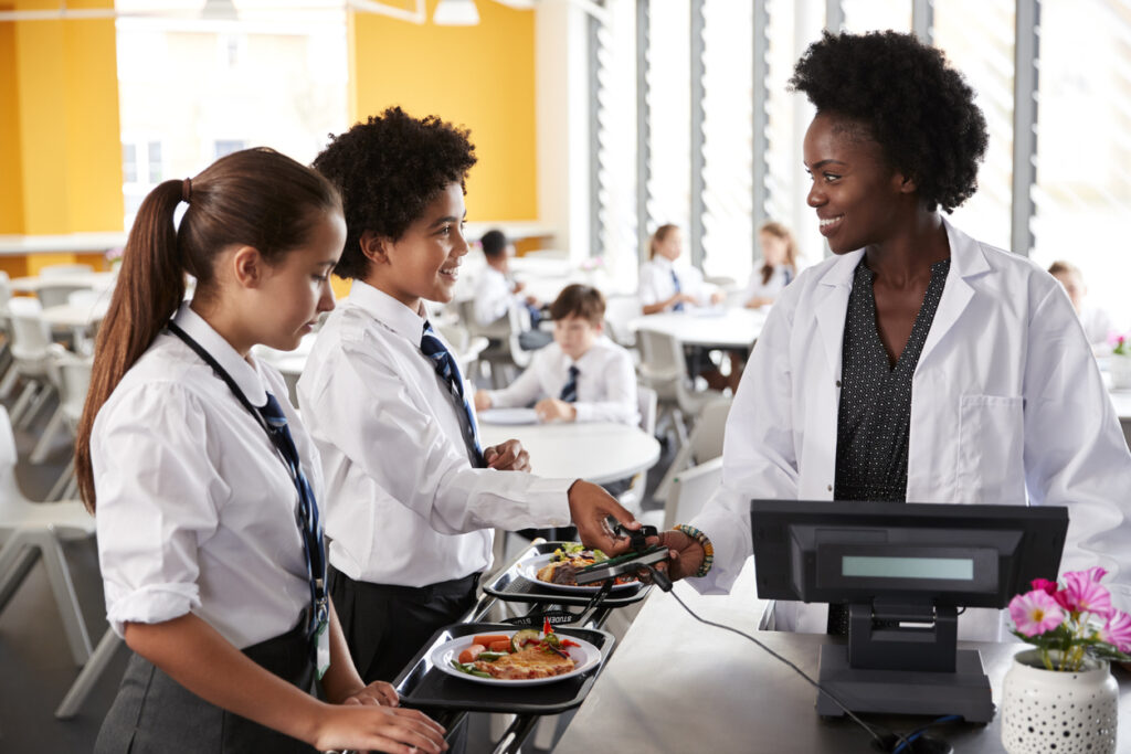 Two students in school uniforms are engaging with a member of the cafeteria staff at a checkout counter equipped with an advanced EPOS system. The students are holding trays with meals, while the bright, bustling cafeteria is filled with other students in the background.