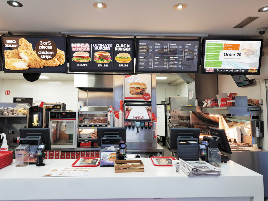 Fast-food restaurant counter featuring EPOS systems beneath menu screens displaying BBQ sauce, chicken strips, burgers, and combination meals such as the Mega Burger and Cluck Burger. An order number 20 flashes on screen above two cash registers on the white countertop.
