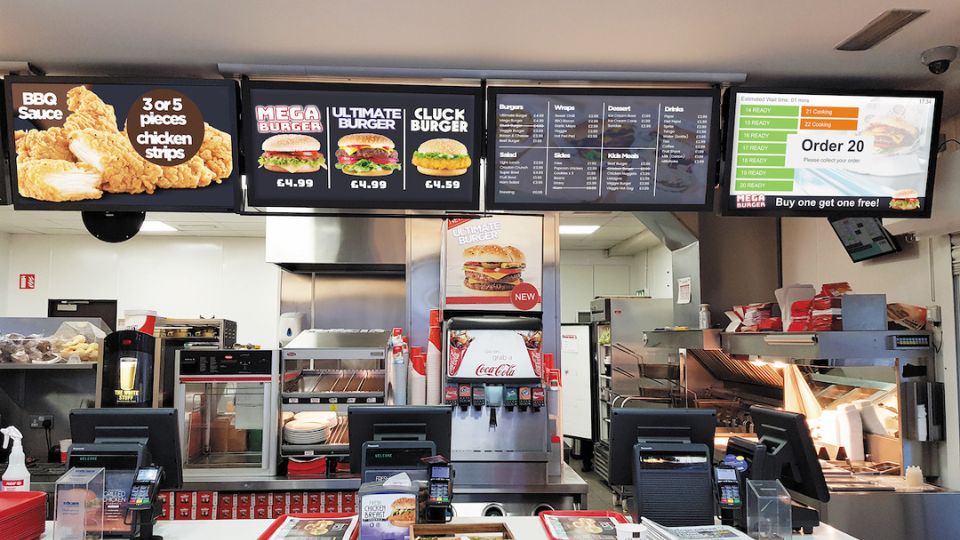 A fast food restaurant counter features digital menu boards showcasing BBQ chicken strips, burgers, and meal deals. With EPOS systems for seamless ordering, cooking equipment, and promotional posters in view, the bright interior ensures that menu prices are easily accessible.