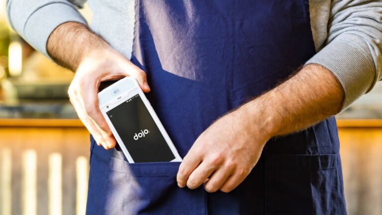 A person in a blue apron holds a white card machine displaying dojo on the screen, improving service efficiency in cafés, pubs, and restaurants.