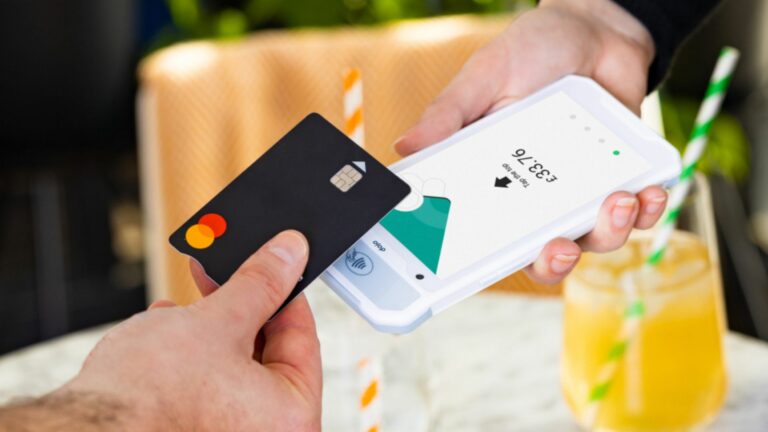 In a bustling café, someone holds a sleek black credit card, ready to tap against the white payment terminal presented by another. A glass with a yellow drink and straw sits on the table, highlighting the efficient service in this vibrant setting.