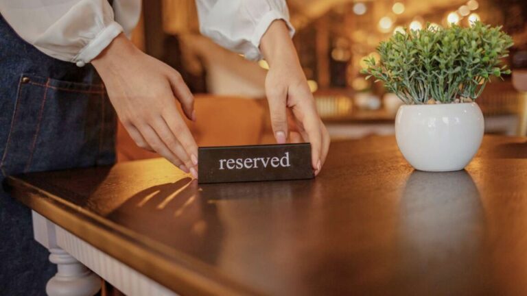 Hands delicately place a reserved sign on the wooden table in a warmly lit restaurant, where a small potted plant adds a touch of greenery. Nearby, the glow from EPOS systems subtly enhances the inviting ambience.