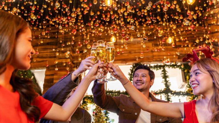 A group of people in festive attire clink champagne glasses in a celebratory toast, surrounded by holiday decorations such as fairy lights and baubles. Amidst the joy, they express excitement over increased Christmas profits. Smiles all around capture this joyful, festive moment.