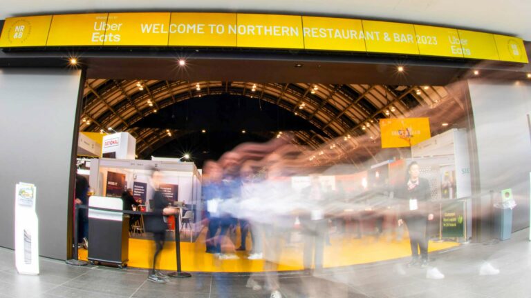 Blurred figures walk past the entrance to the Northern Restaurant & Bar 2024 event. A large yellow banner featuring the Uber Eats logo stands prominently. Inside, various restaurant stands and a curved ceiling welcome attendees.