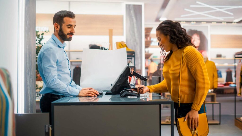 A woman in a yellow jumper, holding a matching handbag, uses the cutting-edge EPOS system at the shop counter. A man in a blue shirt stands behind the counter, smiling warmly. The shops modern interior is visible in the background.