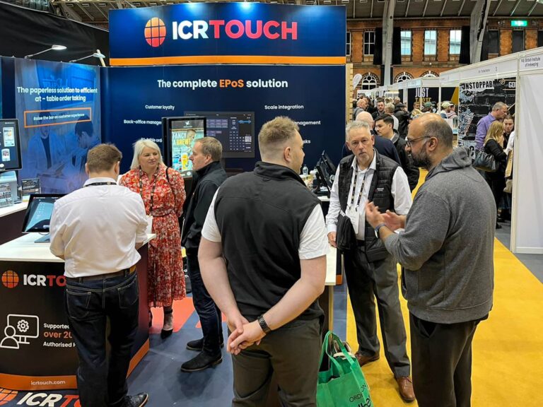 A group of people stand and converse at an exhibition stand with the banner ICRTouch promoting EPoS solutions. The area is bustling with attendees, and digital displays are visible in the background.