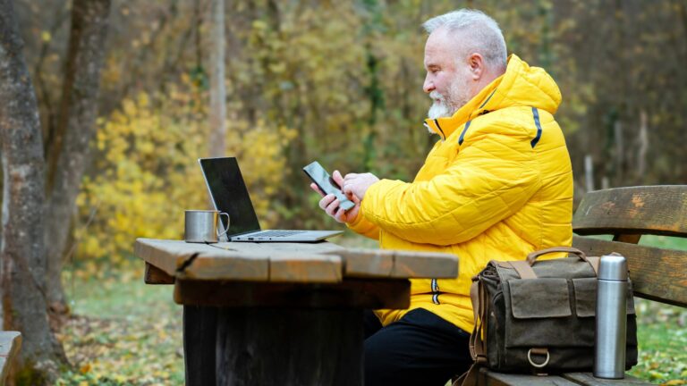A person with a grey beard wearing a bright yellow jacket sits at a wooden picnic table outdoors, using a smartphone. With a laptop, metal mug, and bag on the table, they are managing businesses remotely against the backdrop of trees adorned with autumn foliage.