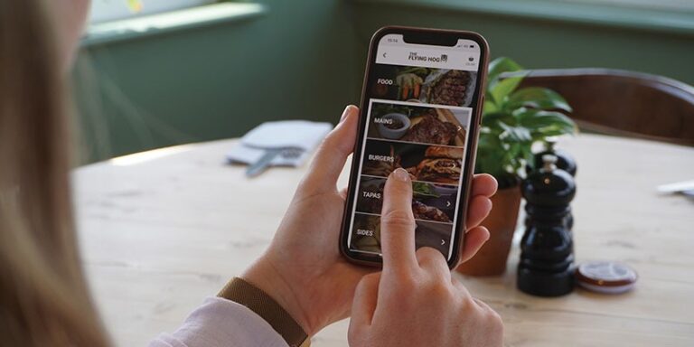 An individual is holding a smartphone, browsing a menu app powered by EPOS software, with options such as main courses, burgers, tapas, and side dishes. The phone is held in one hand while the other taps the screen. In the background are a table with a plant, salt and pepper shakers, and a chair.