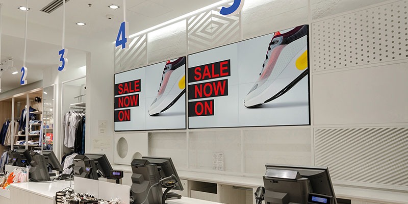Store interior with checkout counters equipped with EPOS software and digital screens displaying Sale Now On over a trainer image. Numbered signs hang from the ceiling above the counters, and clothing items are visible on the left side.