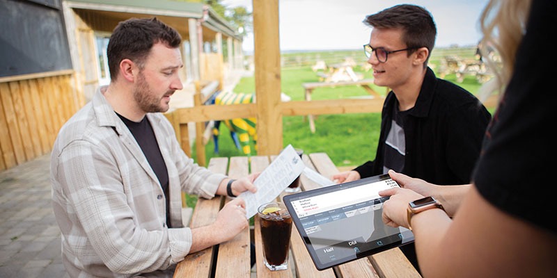 Two men seated at an outdoor wooden table look over a menu. One holds a drink as a nearby person uses EPOS software on their tablet to take the order. The scene takes place in a garden area with picnic tables and lush grass in the background, creating a relaxed dining atmosphere.