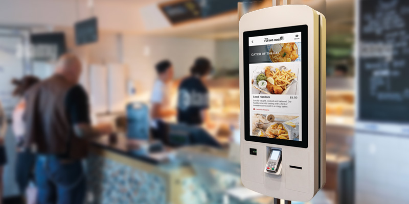 A digital ordering kiosk, integrated with EPOS Software, displays a menu with food items and prices. Several people are in the background, standing at the counter. The kiosk includes a card payment terminal for smooth transactions.