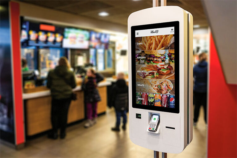 A self-service kiosk equipped by CCR Systems in a fast-food restaurant showcases menu items like chips, burgers and wraps. A payment terminal is integrated below the screen. In the background, customers line up at the counter to order food.