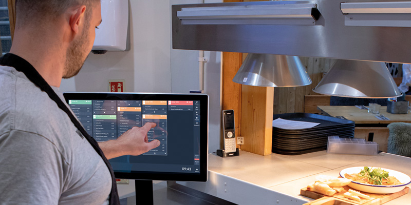 In a bustling kitchen, a person in an apron expertly navigates EPOS software on a touchscreen monitor. Plates of food grace the counter in the background, accompanied by a stack of trays and sleek metal hanging lamps that add to the culinary ambience.