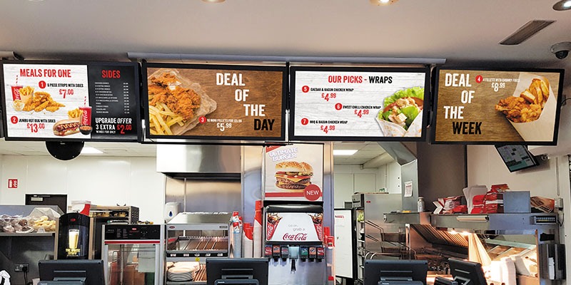 Fast food restaurant interior featuring digital menu boards powered by EPOS software, displaying various meals and deals. Offerings include Deal of the Day, Our Picks - Wraps, Deal of the Week, and Meals for One with prices. Kitchen area visible in the background.