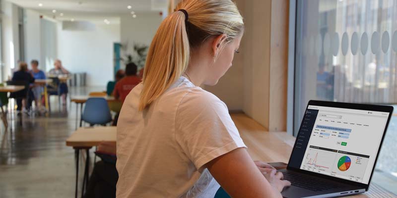 A woman with blonde hair tied back is working on a laptop at a high table by a window. The screen displays charts and data, likely linked to EPOS Software. In the background, people sit at tables in a well-lit, modern café setting.