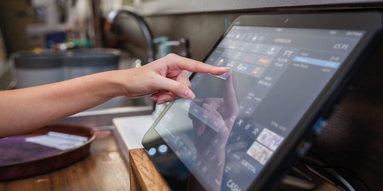 A hand using a point-of-sale touchscreen system in a retail or restaurant setting effortlessly navigates the EPOS software. The screen displays various buttons and options, indicating a transaction or order process. The environment has a modern and organised appearance.