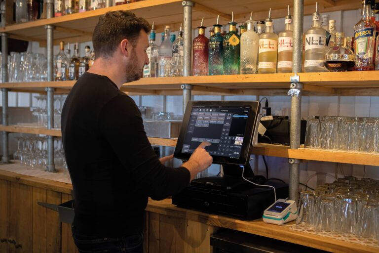 A person in a black shirt smoothly navigates the ICRTouch point-of-sale system at a bar. Shelves lined with diverse bottles of liquor and elegant glassware provide a New Look to the scene. A Dojo card reader sits conveniently beside the sleek terminal.