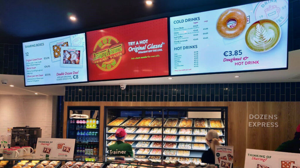 A Krispy Kreme store greets customers with vibrant digital signage showcasing a menu of hot drinks and doughnut deals, including a tempting offer for a glazed doughnut and hot drink combo. Behind the counter, staff serve eager patrons with an array of delicious doughnuts in the background.