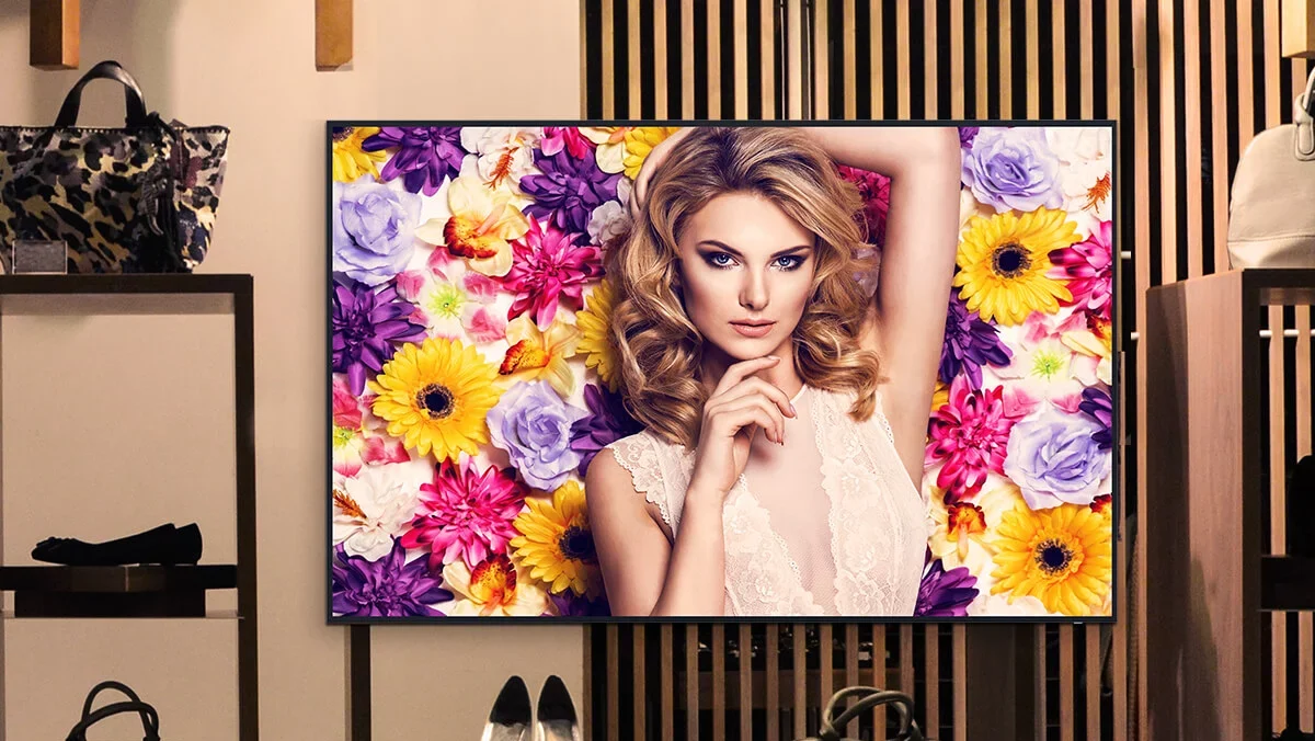 A digital signage screen features a fashionable model with curly hair set against a vibrant floral backdrop. The scene unfolds in a stylish retail space, adorned with shelves displaying chic handbags and shoes.