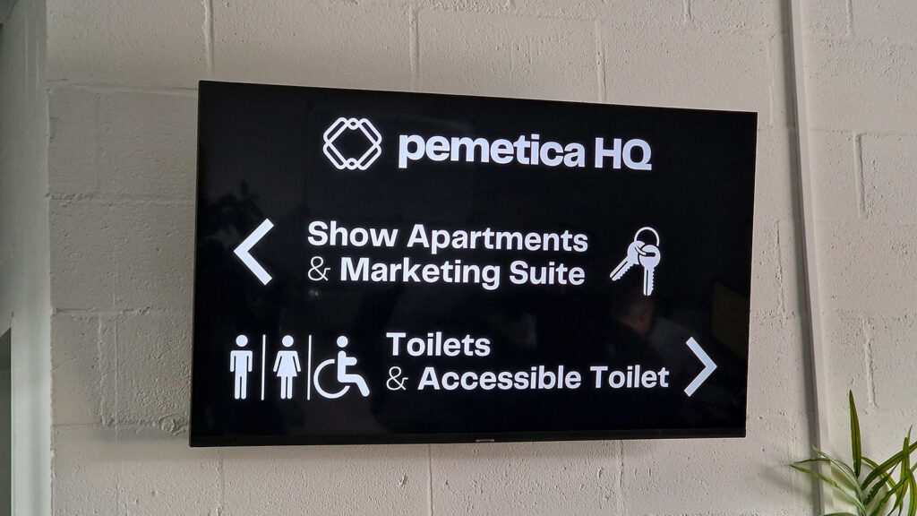 At pemetica HQ, a digital signage system enhances wayfinding with arrows directing visitors to Show Apartments & Marketing Suite and Toilets & Accessible Toilet, complete with icons for gendered restrooms and accessibility.