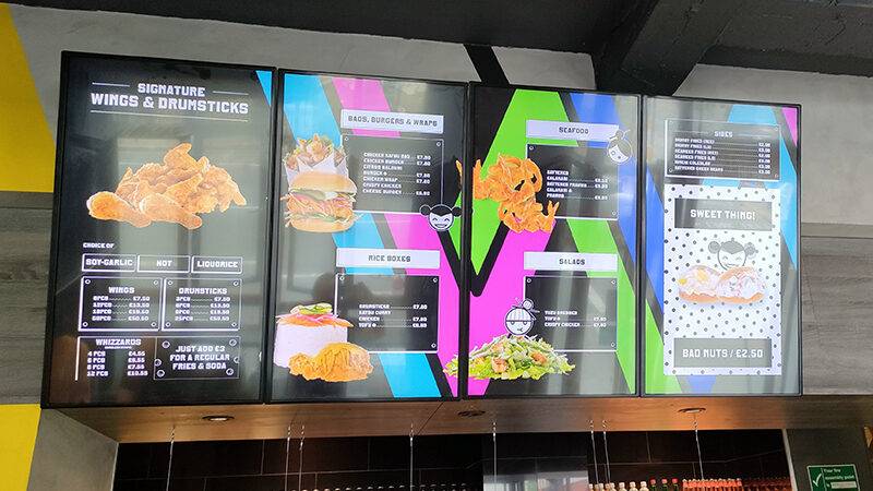 A vibrant digital menu board showcases colourful sections for chicken wings & drumsticks, burgers & wraps, rice boxes, seafood, sides and desserts. Each section features images and prices. The playful design uses bright colours and engaging graphics to captivate attention.