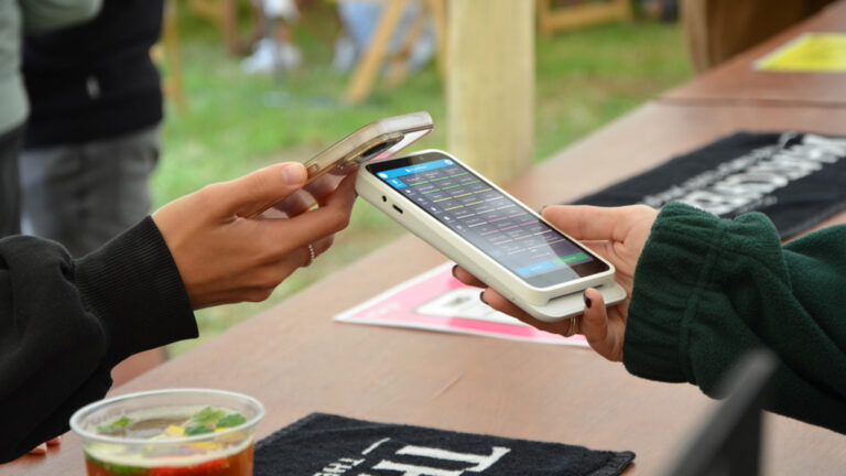 Two people are engaging in a mobile payment transaction outdoors using TouchPoint Lite. One person holds a smartphone, and the other holds a payment device. A drink in a transparent cup and a black cloth lie casually on the wooden table nearby, completing the scene.