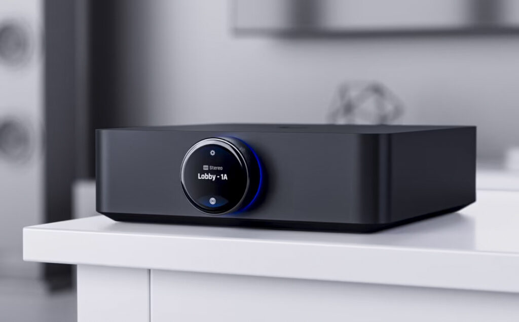 A sleek black electronic device, the UniFi PowerAmp, features a circular digital display on the front showing the text Lobby - 1A. It rests on a white surface against a modern, blurred backdrop.