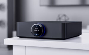 A sleek black electronic device, the UniFi PowerAmp, features a circular digital display on the front showing the text Lobby - 1A. It rests on a white surface against a modern, blurred backdrop.