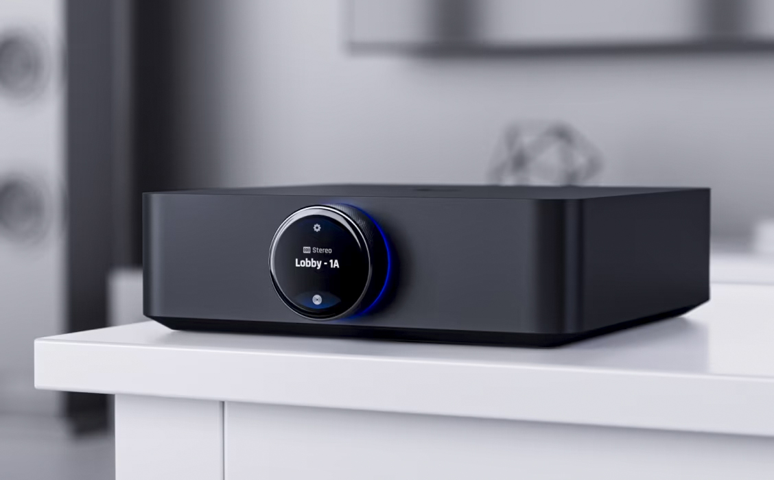 A sleek black electronic device, the UniFi PowerAmp, features a circular digital display on the front showing the text Lobby - 1A. It rests on a white surface against a modern, blurred backdrop.